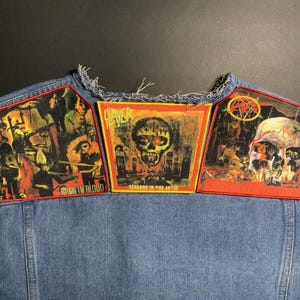 May include: A denim vest with three Slayer band patches. The patches feature album art with skulls and the band's logo. The patches are red, orange, and yellow with black and green accents.