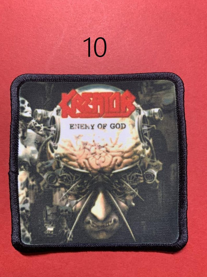 Peut inclure: &Eacute;cusson carr&eacute; avec le nom du groupe "KREATOR" en lettres rouges au-dessus du texte "ENEMY OF GOD". L'&eacute;cusson pr&eacute;sente un graphisme d&eacute;taill&eacute; d'un visage m&eacute;canique avec un cerveau et des composants m&eacute;talliques. L'&eacute;cusson a une bordure noire.