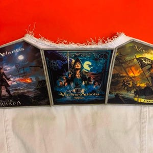 Op de afbeelding: Drie rechthoekige stoffen patches met Visions of Atlantis albumillustraties, waaronder Pirates en Armada, bevestigd op een wit spijkergilet. De patches tonen nautische en fantasie-scènes met levendige kleuren, tekst en gerafelde randen.
