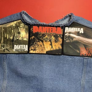 Könnte beinhalten: Eine blaue Jeansweste mit drei rechteckigen Aufnähern der Band Pantera. Die Aufnäher zeigen Bilder und Text, darunter den Bandnamen und Albumtitel. Die Weste hat ein abgenutztes Aussehen.