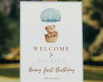 May include: A welcome sign on a wooden easel. The sign features a watercolor illustration of a teddy bear in a hot air balloon. Text reads "Welcome to Noah's Beary first Birthday" and the date 08.19.2025.