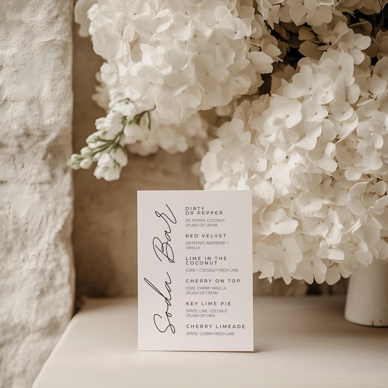 Op de afbeelding: Een witte Soda Bar menukaart met zwarte scripttekst met frisdranksmaken zoals Cherry on Top en Key Lime Pie. De kaart staat tegen een achtergrond van witte bloemen en een wit oppervlak.
