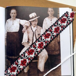 May include: A beaded bookmark with a floral pattern in red, white, and black. The bookmark is placed over an open book with a sepia-toned photograph of three people. A white cord is attached to the bookmark.