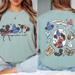 May include: A light green t-shirt featuring Disney-themed graphics. The front displays Mickey Mouse ears, a wizard hat, and Minnie Mouse ears with the text "Some imagination, huh?" The back showcases Mickey as a wizard, tickets, and other themed elements.