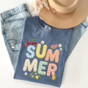 May include: A blue t-shirt with the word "SUMMER" in colorful, patchwork-style letters. The shirt features embroidered fruit and flower details. A pair of light-wash jeans and a tan hat are also in the image.
