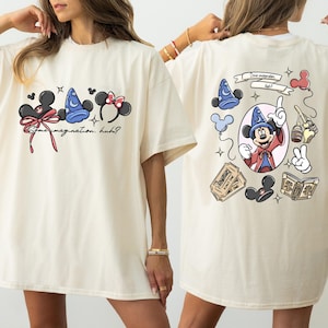 May include: Cream-colored t-shirt featuring Disney-themed graphics. The front displays Mickey Mouse ears, a sorcerer's hat, and text. The back showcases Mickey Mouse as a sorcerer, along with other Disney elements.