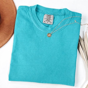 May include: A solid turquoise short-sleeve t-shirt with a crew neckline. The inner neck label reads Comfort Colors, 100% Cotton, Made in Honduras, S P CH. A thin gold-toned chain necklace with a small circular pendant rests on the fabric.
