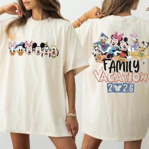 May include: Cream-colored t-shirt featuring a colorful graphic of Disney characters, including Mickey Mouse, Minnie Mouse, Donald Duck, and Goofy. The back of the shirt displays the text "Family Vacation 2026" with additional Disney characters.