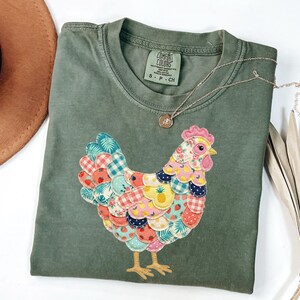 May include: Olive green t-shirt featuring a colorful patchwork chicken design. The chicken has a pink comb and wattle, with a variety of patterned fabrics in its feathers. A gold necklace with a pendant is draped on the shirt.