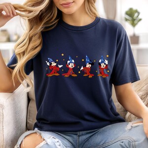 May include: Navy blue t-shirt featuring a graphic of Mickey Mouse in a wizard costume. The design shows Mickey in a red robe and blue wizard hat with stars. A comfortable, casual tee for Disney fans.