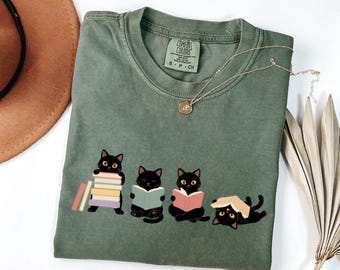 May include: An olive green crew neck t-shirt featuring a graphic of four black cats interacting with books. One cat holds a stack of books, two cats are reading open books, and one cat lies on its back with a book resting on its head. The shirt has a Comfort Colors brand label.