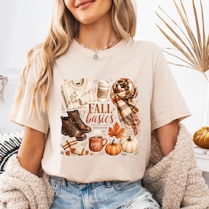 May include: A cream-colored t-shirt featuring a graphic print of autumn items including a sweater, boots, a scarf, a coffee cup, a mug, pumpkins, and leaves. The text "FALL basics" and "autumn essentials" is printed in the center of the design.