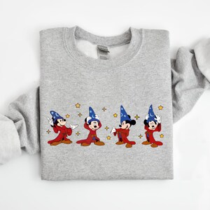 May include: A light gray sweatshirt featuring an embroidered design of Mickey Mouse in a wizard costume. The design includes four Mickey Mouse figures in red robes and blue wizard hats, with gold stars scattered around them. The sweatshirt has long sleeves.