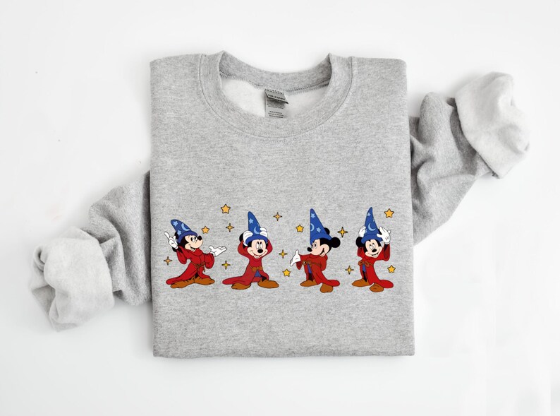 May include: A light gray sweatshirt featuring an embroidered design of Mickey Mouse in a wizard costume. The design includes four Mickey Mouse figures in red robes and blue wizard hats, with gold stars scattered around them. The sweatshirt has long sleeves.