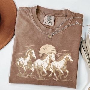 May include: A brown short-sleeve t-shirt featuring a graphic print of three horses running in front of a large sun. The inner neck label reads Comfort Colors, Heavyweight Garment Dye, Ring Spun, 100% Cotton, Made in Honduras, S-P-CH.