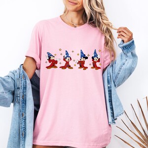 May include: A light pink t-shirt featuring a graphic of Mickey Mouse in a wizard costume. Mickey is wearing a blue wizard hat and red robe. The graphic includes gold stars. The shirt has short sleeves and a relaxed fit.