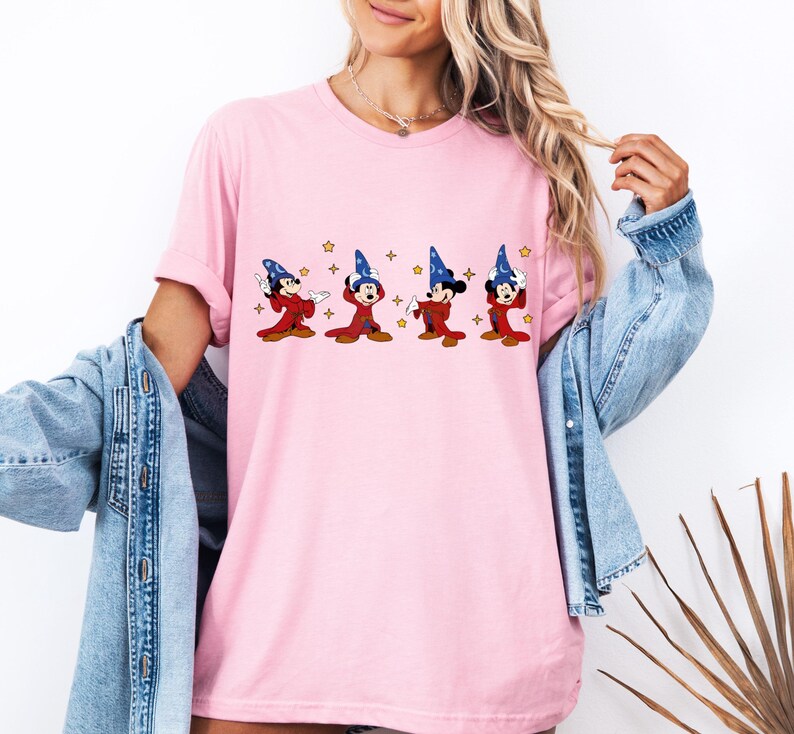 May include: A light pink t-shirt featuring a graphic of Mickey Mouse in a wizard costume. Mickey is wearing a blue wizard hat and red robe. The graphic includes gold stars. The shirt has short sleeves and a relaxed fit.