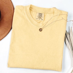 May include: A solid light yellow short-sleeve t-shirt with a crew neckline. The inner neck label reads Comfort Colors, 100% cotton, made in Honduras, size S P CH. A thin gold-toned chain necklace with a small circular pendant rests on the fabric.