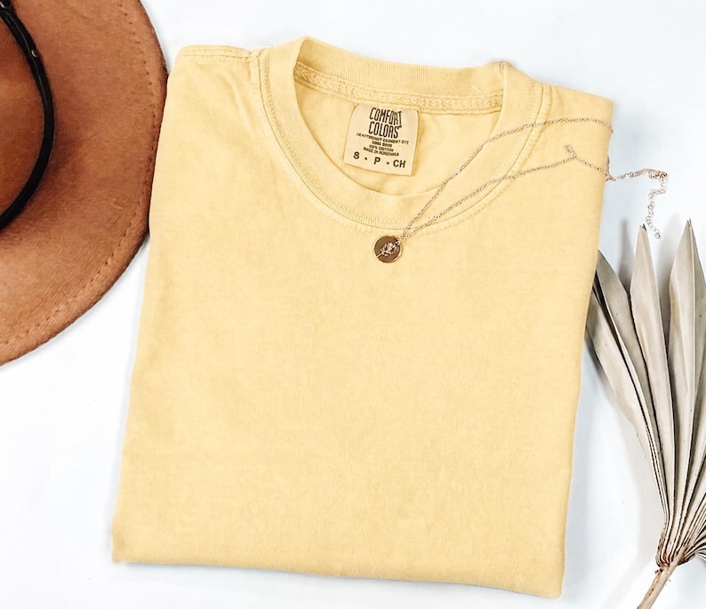 May include: A solid light yellow short-sleeve t-shirt with a crew neckline. The inner neck label reads Comfort Colors, 100% cotton, made in Honduras, size S P CH. A thin gold-toned chain necklace with a small circular pendant rests on the fabric.