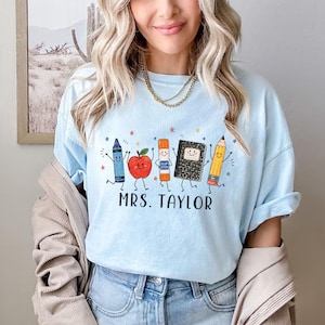 May include: A light blue t-shirt featuring a graphic of a blue crayon, a red apple, an orange glue stick, a black composition notebook, and a yellow pencil, all with smiling faces. The text "MRS. TAYLOR" is printed in black below the characters.
