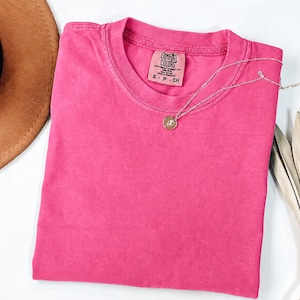 May include: A solid hot pink short-sleeve t-shirt with a crew neckline. The inner neck label reads Comfort Colors, 100% cotton, made in Honduras, size S. A thin silver-toned chain necklace with a small circular pendant rests on the fabric.
