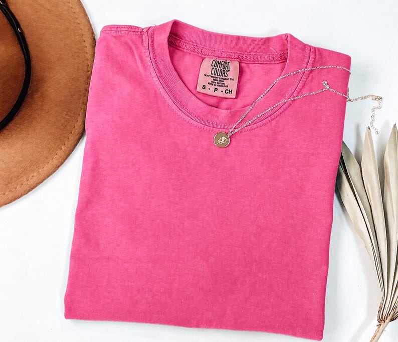 May include: A solid hot pink short-sleeve t-shirt with a crew neckline. The inner neck label reads Comfort Colors, 100% cotton, made in Honduras, size S. A thin silver-toned chain necklace with a small circular pendant rests on the fabric.