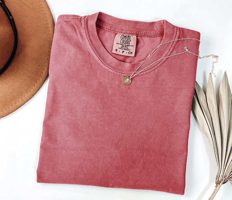 May include: A dusty rose-colored short-sleeve t-shirt with a crew neckline. The interior neck label reads Comfort Colors, Heavyweight Garment Dye, 100% Cotton, Made in Honduras, S P CH. A thin gold-toned chain necklace with a small circular pendant rests on top.