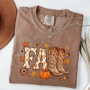 May include: A brown short-sleeve t-shirt featuring a graphic print that spells the word FALL. The letters are decorated with a checkered pattern, cactus silhouettes, and leopard print cowboy boots. A pumpkin, maple leaves, and acorns surround the text.
