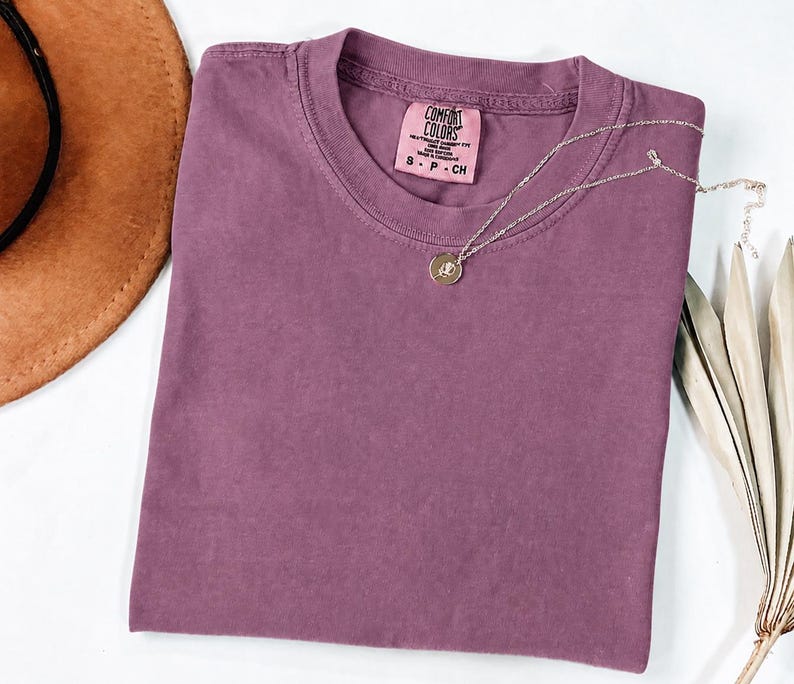 May include: A solid mauve short-sleeve t-shirt with a crew neckline and a Comfort Colors brand label inside the collar. A delicate silver-toned chain necklace with a small circular pendant featuring a floral engraving rests on the fabric.