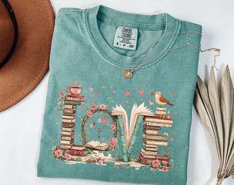 May include: Teal Comfort Colors t-shirt with a floral and book-themed design spelling "LOVE". The design includes stacks of books, a teacup, a bird, and a floral wreath. A silver necklace with a pendant is on the shirt.