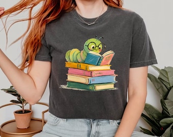 May include: A charcoal gray short-sleeve t-shirt featuring a graphic illustration of a green caterpillar wearing black-rimmed glasses while reading an open book. The caterpillar rests on a stack of four colorful books in yellow, pink, blue, and green.