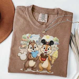 May include: A brown Comfort Colors t-shirt featuring a graphic of Chip and Dale holding treats in front of a theme park scene. The shirt has a crew neck and short sleeves. A silver necklace with a small pendant is draped across the shirt.