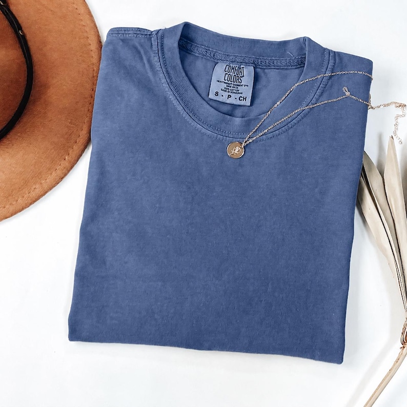 May include: A solid blue short-sleeve t-shirt with a crew neckline and a Comfort Colors brand label printed on the inside collar. A thin gold-toned chain necklace with a small circular pendant rests on the center of the fabric.