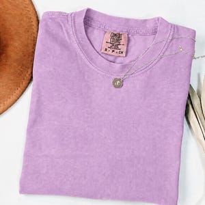 May include: A solid light purple short-sleeve t-shirt with a crew neckline. The inner neck label reads Comfort Colors, Heavyweight Garment Dye, 100% Cotton, Made in Honduras, S P CH. A thin silver-toned chain necklace with a small circular pendant rests on top.
