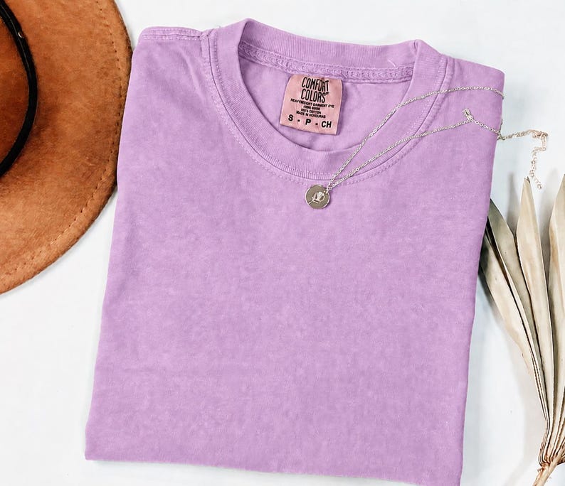 May include: A solid light purple short-sleeve t-shirt with a crew neckline. The inner neck label reads Comfort Colors, Heavyweight Garment Dye, 100% Cotton, Made in Honduras, S P CH. A thin silver-toned chain necklace with a small circular pendant rests on top.
