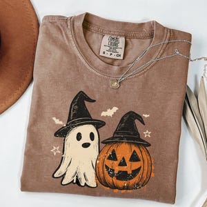 May include: A brown t-shirt featuring a graphic of a ghost and a jack-o'-lantern, both wearing black witch hats. The design includes small bats and stars. The shirt has a Comfort Colors label at the neck and is paired with a silver chain necklace.