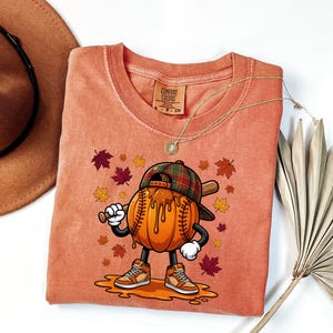 May include: A terracotta-colored t-shirt featuring a graphic of a cartoon pumpkin wearing a plaid baseball cap and sneakers, holding a baseball bat. The pumpkin is surrounded by falling autumn leaves, with the Comfort Colors brand label visible.