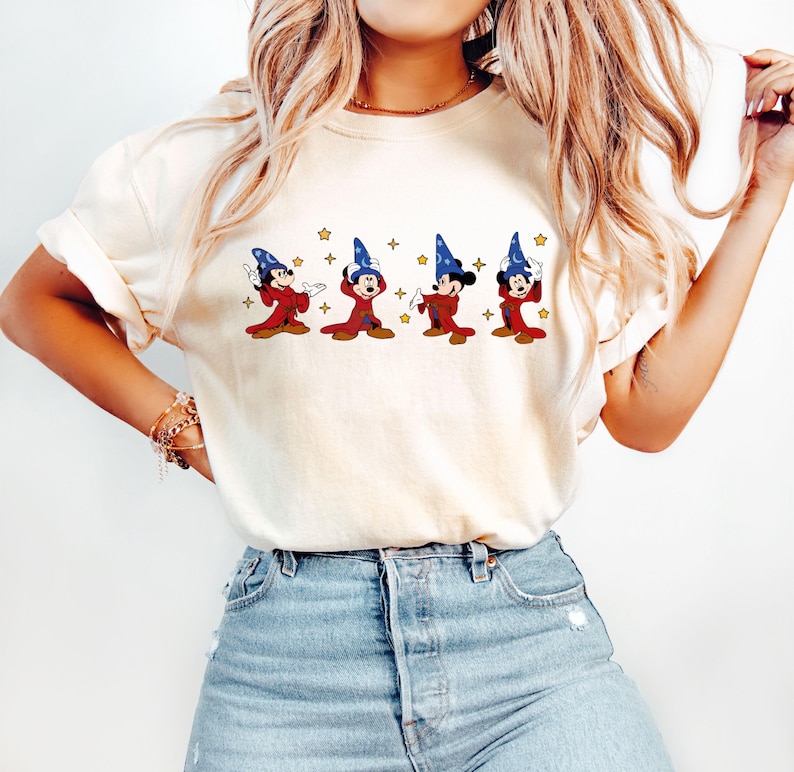 May include: Cream-colored t-shirt featuring a graphic of Mickey Mouse as a sorcerer in a red robe and blue hat. The design includes gold stars. The shirt is paired with blue jeans.