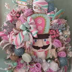 May include: A pink and mint green Christmas wreath featuring a nutcracker face, candy canes, and snowflake ornaments.