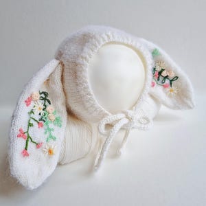 Puede incluir: Un gorro de bebé blanco tejido con largas orejas de conejo. Las orejas están adornadas con flores bordadas en rosa, verde y amarillo. Un lazo blanco asegura el gorro bajo la barbilla. El gorro está diseñado para un bebé.