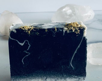 Puede incluir: Una pastilla rectangular de jabón hecho a mano. El jabón es principalmente negro con un borde inferior veteado blanco y cubierto con hierbas secas. Objetos cristalinos blancos en el fondo.
