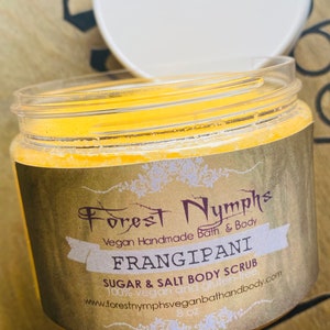 Puede incluir: Un tarro transparente de exfoliante corporal de az&uacute;car y sal Frangipani con tapa blanca. La etiqueta dice "Forest Nymphs" y "Vegan Handmade Bath & Body". El tarro contiene 227 gramos de producto.