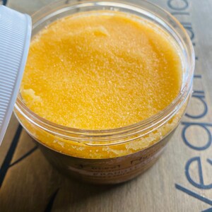 Puede incluir: Un tarro de pl&aacute;stico transparente lleno de un exfoliante texturizado de color amarillo-naranja. La tapa est&aacute; parcialmente abierta, revelando el producto en su interior. El tarro est&aacute; etiquetado con texto que incluye las palabras "Forest Nymph" y "Frankincense & Salt".
