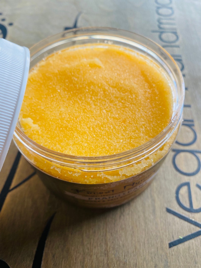 Puede incluir: Un tarro de pl&aacute;stico transparente lleno de un exfoliante texturizado de color amarillo-naranja. La tapa est&aacute; parcialmente abierta, revelando el producto en su interior. El tarro est&aacute; etiquetado con texto que incluye las palabras "Forest Nymph" y "Frankincense & Salt".