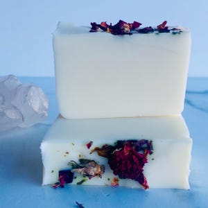 May include: Two white bars of soap with dried rose petals and green herbs on top. The soap is on a light blue surface.