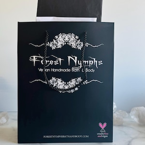 Puede incluir: Bolsa de la compra negra con texto blanco y diseño floral. La bolsa dice "Forest Nymphs Vegan Handmade Bath & Body" y "cruelty-free and vegan". La bolsa tiene asas de cuerda negra y está sobre una superficie de mármol.