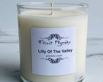 Puede incluir: Un tarro de vela de vidrio transparente lleno de cera blanquecina. Una etiqueta blanca con el texto "Forest Nymphs" y "Lilly Of The Valley" está centrada en el tarro. La vela tiene una sola mecha y está etiquetada como una vela de piedras preciosas.