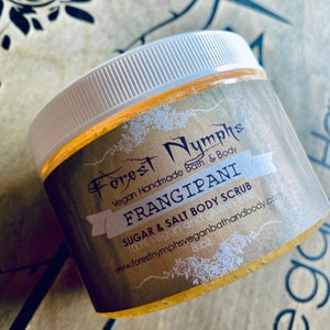 Puede incluir: Un tarro de exfoliante corporal de az&uacute;car y sal Frangipani de Forest Nymphs. El tarro tiene una tapa blanca y una etiqueta dorada con el nombre y los detalles del producto. El exfoliante es de 227g y vegano.