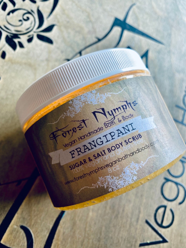 Puede incluir: Un tarro de exfoliante corporal de az&uacute;car y sal Frangipani de Forest Nymphs. El tarro tiene una tapa blanca y una etiqueta dorada con el nombre y los detalles del producto. El exfoliante es de 227g y vegano.