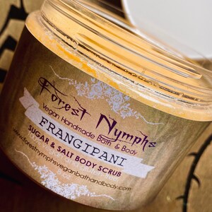 Puede incluir: Un tarro transparente de exfoliante corporal de az&uacute;car y sal de Frangipani con tapa blanca. La etiqueta dice "Forest Nymphs Vegan Handmade Bath & Body". El tarro contiene un exfoliante amarillo-anaranjado. La etiqueta es marr&oacute;n con texto blanco y morado.